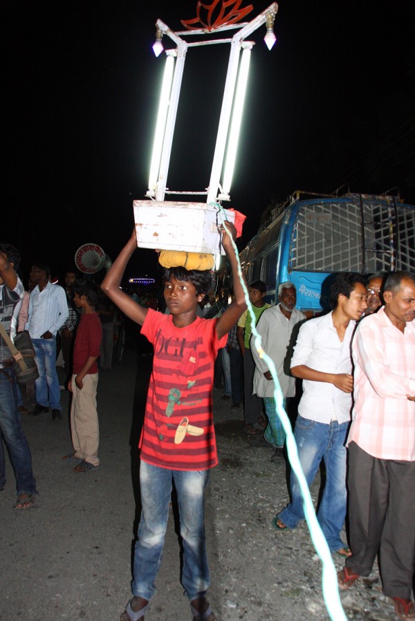 While male relatives of a wedding couple dance and celebrate on the road, a young girl leads the way with large fluorescent lights about her head.